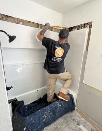 A person in a black T-shirt and beige pants is measuring a gap above a white tiled bathtub. They stand on a protective cover placed over the tub. Surrounding walls are partially stripped.